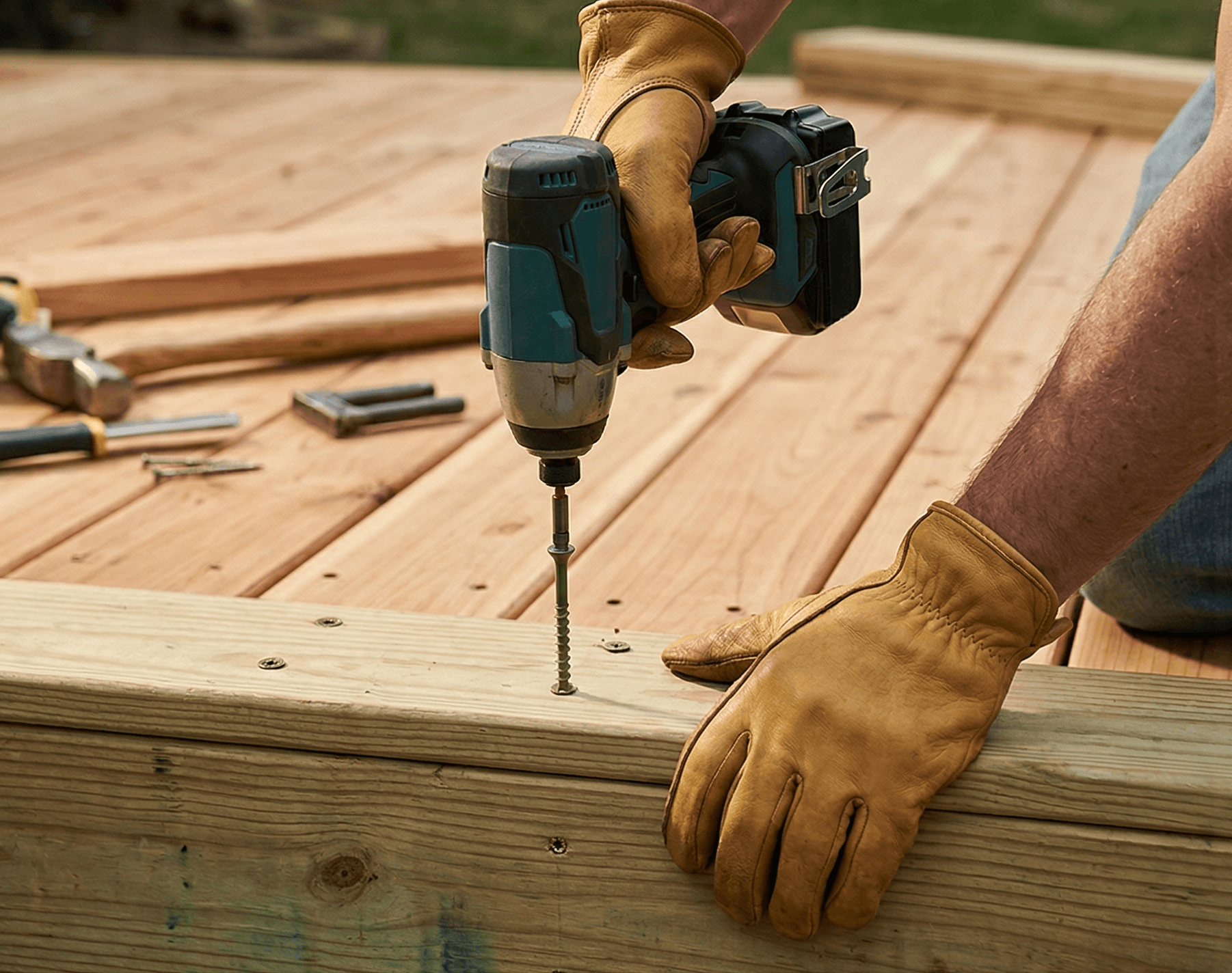 Person wearing leather work gloves driving a deck screw into a wood board with a cordless drill during deck construction