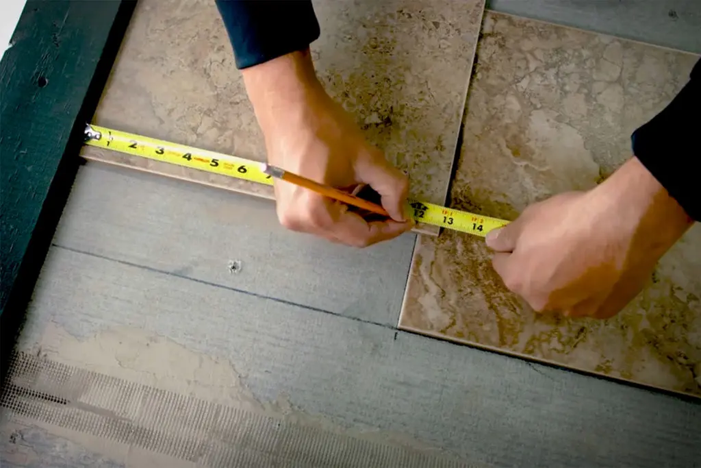 Hands measuring and marking a stone floor tile with a tape measure and pencil before cutting during a tile installation